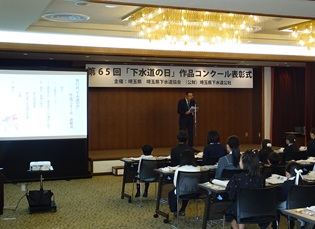 舞台上で説明する職員と聞く受賞者約２０名